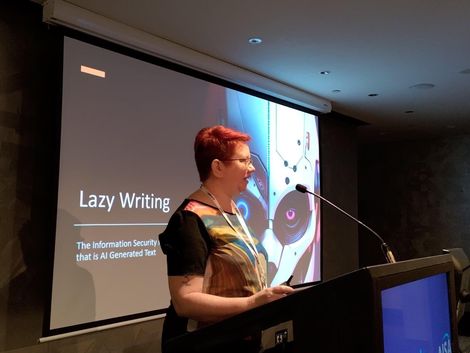 Speaker Kristine Sihto stands in front of her title slide - Lazy Writing: The information security nightmare that is AI generated text. The image beside it is an AI generated image of a cyborg's face, half in shadow. It is constructed of white plastic, with one blue eye set into metal and one pink eye in an almost reptilian black surrounding.